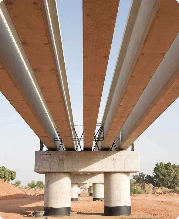 Low-angle view looking up beneath a newly constructed concrete viaduct, showing heavy precast bridge girders supported by large cylindrical concrete piers.