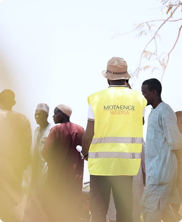 Rear view of a Mota-Engil Nigeria site engineer wearing a high-visibility yellow vest and bucket hat, standing outdoors and engaging with local community members.