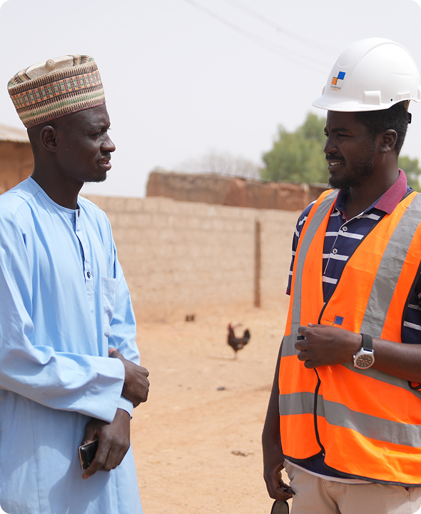 A Mota-Engil Nigeria site engineer wearing an orange safety vest and white hard hat speaking warmly with a local community member in traditional blue attire.