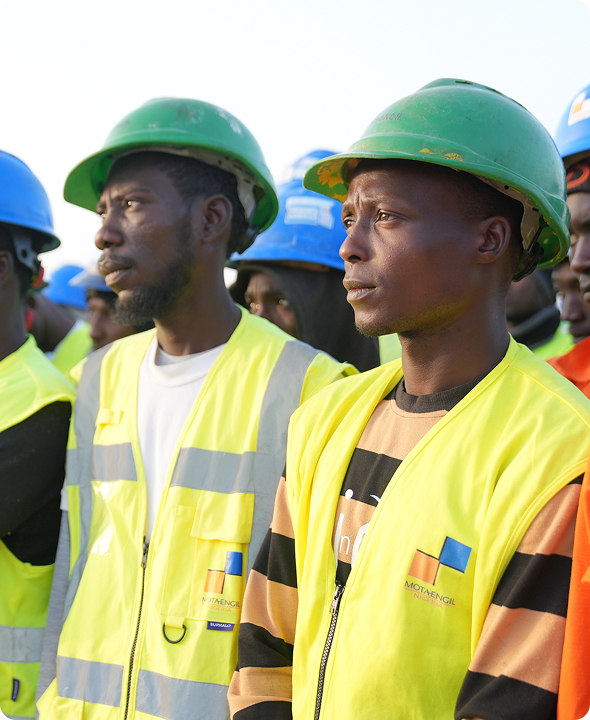Mota-Engil Nigeria construction workers wearing green hard hats and high-visibility yellow safety vests standing together on-site.