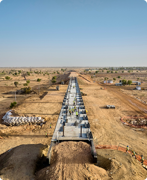 High-angle perspective view of a long concrete bridge deck under construction, with site workers walking along the structure spanning across an arid terrain.