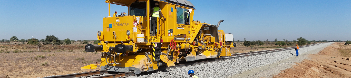 A large yellow track tamping machine operating on a railway line with ballast, while a worker in safety gear inspects the tracks from the side.