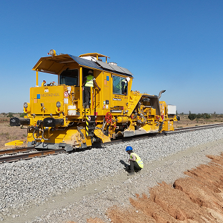 A large yellow track tamping machine operating on a railway line with ballast, while a worker in safety gear inspects the tracks from the side.