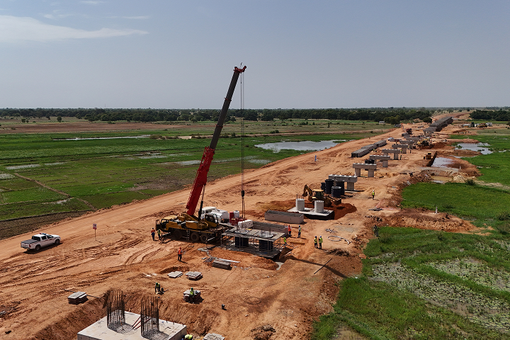 Aerial view of a long infrastructure corridor where a large red mobile crane is positioning heavy elements for concrete bridge piers.