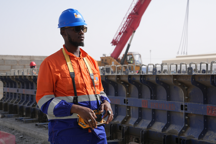 A site worker wearing a blue hard hat, sunglasses, and orange work jacket using a wireless remote control unit with a large red crane in the background.