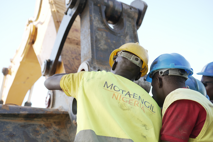 Close-up of two mechanics wearing hard hats and high-visibility vests inspect and repair heavy machinery.
