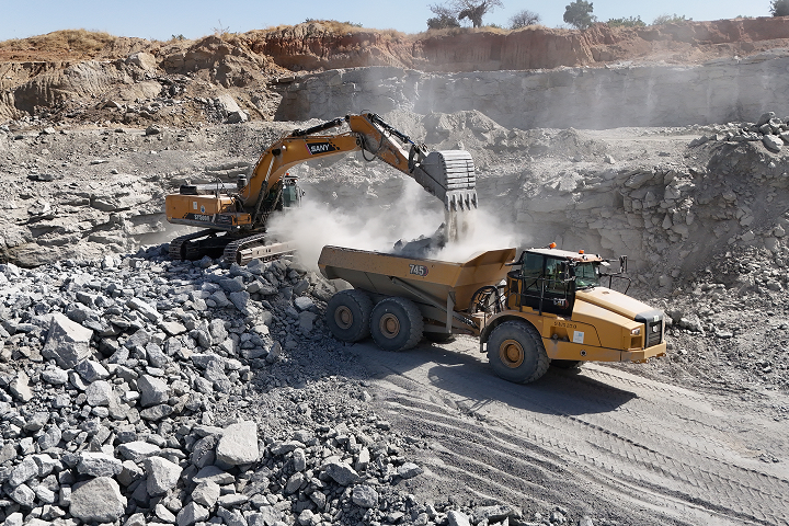 A large excavator loading crushed stone and rock into an articulated yellow dump truck inside a dusty quarry face.