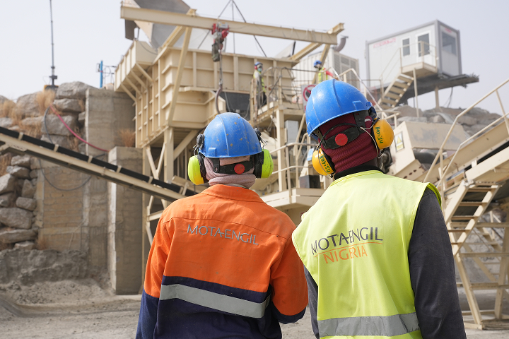 Rear view of two industrial workers wearing protective clothing, ear defenders, and blue hard hats looking toward a large processing plant structure
