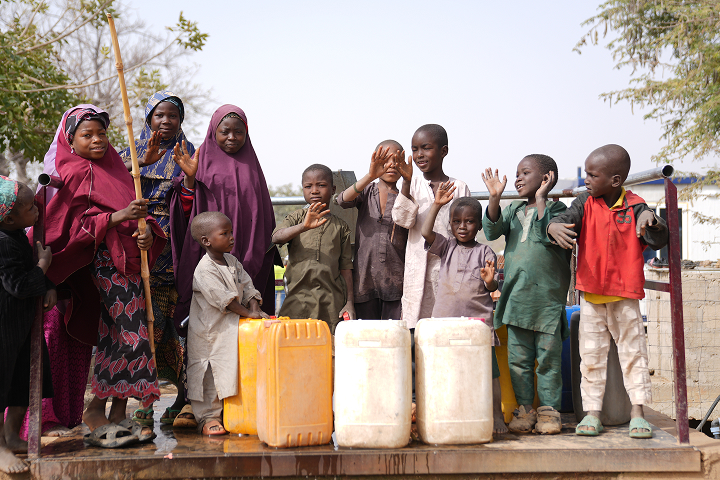 A cheerful group of children and women waving while standing around yellow and white water containers at a clean water access point.