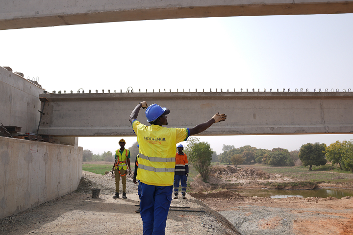 A construction supervisor in a high-visibility yellow vest gesturing upwards near a newly built concrete bridge beam on an infrastructure site.