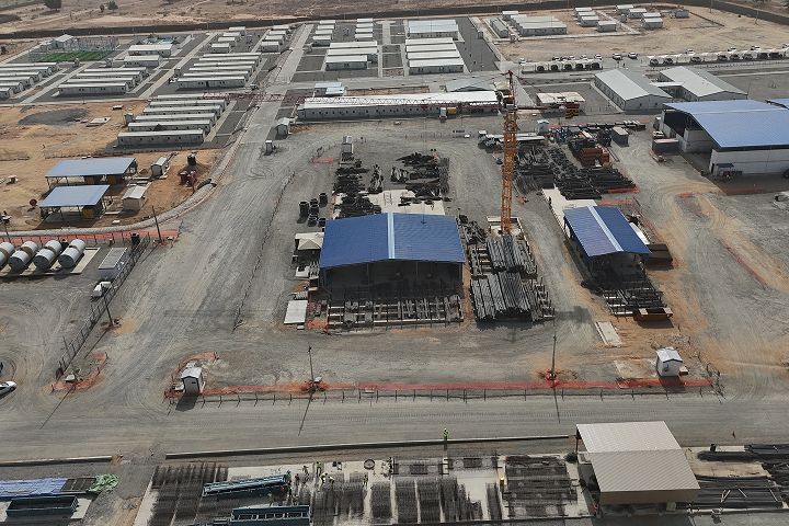 Aerial high-angle view of a vast construction site featuring logistics compounds, modular housing units, materials storage, and a yellow tower crane.