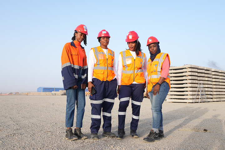 A proud, professional group portrait of four female engineers wearing orange and navy safety jackets, reflective vests, and red hard hats at a construction site with railway sleepers in the background.