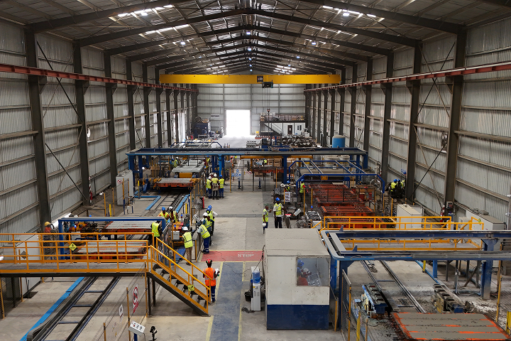 High-angle interior view of a large manufacturing warehouse with overhead yellow gantry cranes, automated assembly lines, and factory technicians at work.