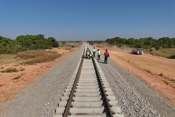 Ground-level view along a new railway track where a crew of construction workers wearing hard hats and high-visibility vests inspect and adjust the rails.