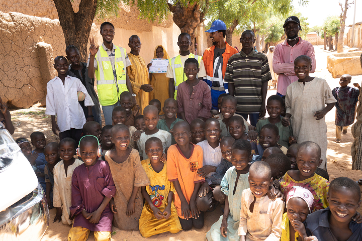 A large, joyful group photo in a Nigerian community. Dozens of smiling children pose in the foreground, with adult community members and Mota-Engil Nigeria staff in reflective vests standing happily behind them.