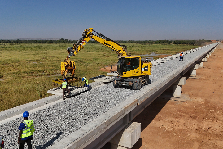 A yellow excavator operating on an elevated concrete railway bridge, placing precast concrete sleepers onto a gravel ballast bed.