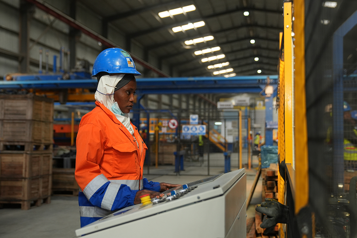 A female factory technician wearing a blue hard hat, white headscarf, and orange work jacket operating an industrial control panel inside a plant.