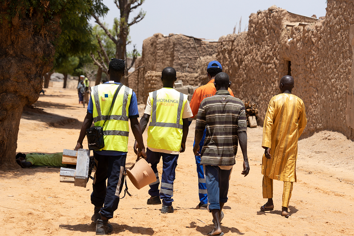 Mota-Engil Nigeria field workers and community members walk together down a sandy road in a rural Nigerian village, carrying an improved cookstove.