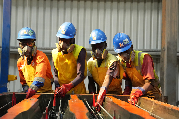 Four factory workers equipped with blue hard hats, protective respirators, and heavy-duty gloves focusing intently on assembling concrete mold components.