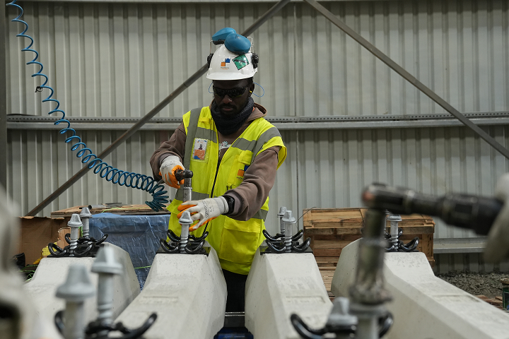 A technician wearing a white hard hat, ear defenders, safety glasses, and a high-visibility vest using a pneumatic tool on concrete railway sleepers.