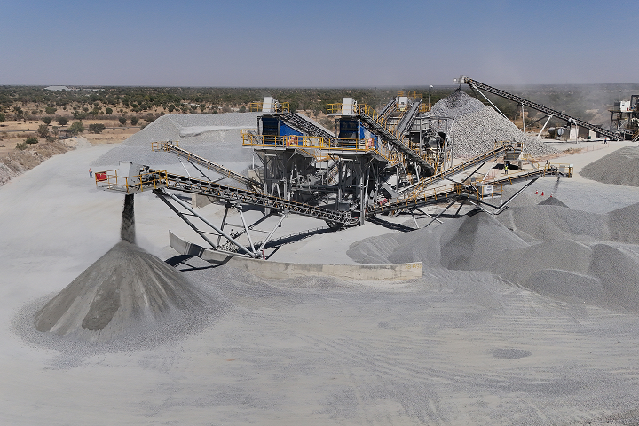 An industrial rock crushing plant with conveyor belts screening aggregates and creating large piles of crushed stone in a quarry yard.