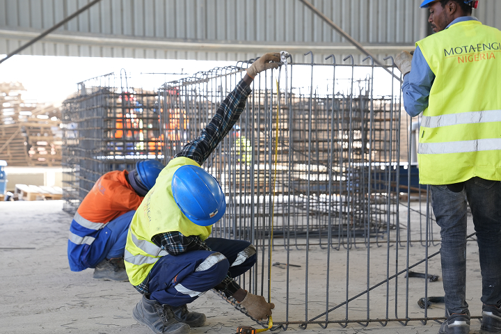 Construction workers wearing blue hard hats and safety gear measuring a rebar steel structure with a tape measure inside a workshop.